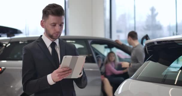 Handsome young Caucasian man standing in car dealership using tablet, looking at camera, smiling, and gesturing yes by shaking head. portrait of confident car dealer at workplace. Cinema 4k ProRes HQ. — 비디오