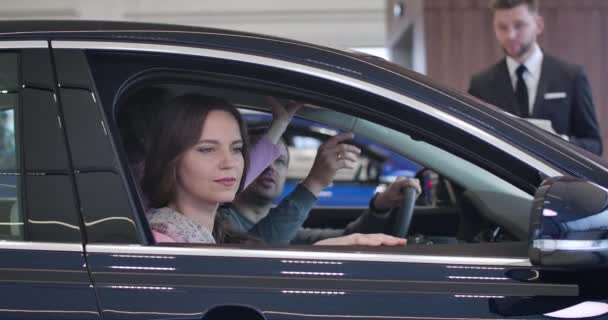 Vista lateral de la joven familia caucásica feliz sentado en el salón de coches y examinar los interiores. Alegre esposa, esposo e hija seleccionando el vehículo en la concesionaria de automóviles. Sede del cine 4k ProRes . — Vídeos de Stock