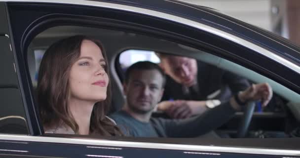 Portrait of charming brunette Caucasian woman looking at car interiors as her husband talking with dealer at the background. Cheerful lady buying automobile with spouse. Cinema 4k ProRes HQ. — 비디오