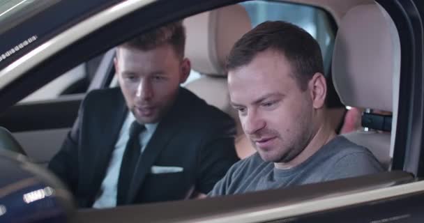 Close-up of Caucasian young man sitting in car salon with blurred dealer at the background. Trader explaining automobile specifications to male customer. Cinema 4k ProRes HQ. — 비디오