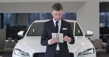 Başarılı genç araba satıcısı elinde tabletle beyaz bir otomobilin önünde kameraya bakıp gülümsüyor. İş yerinde mutlu bir Kafkasyalı tüccar. Sinema 4k Prores Merkezi.