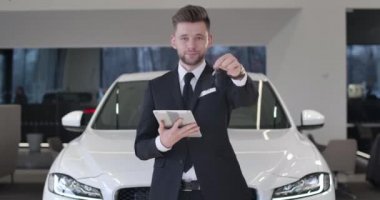 Kameranın anahtarlarını gösteren tabletli yakışıklı beyaz erkek tüccarın portresi. Kendine güvenen genç adam araba galerisinde duruyor ve gülümsüyor. Sinema 4k Prores Merkezi.