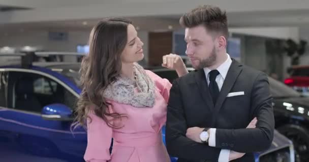 Portrait of happy young Caucasian couple posing in car dealership. Elegant woman in pink dress bragging car keys, handsome man in suit smiling. Automobile industry, joy, success. Cinema 4k ProRes HQ. — Stock Video