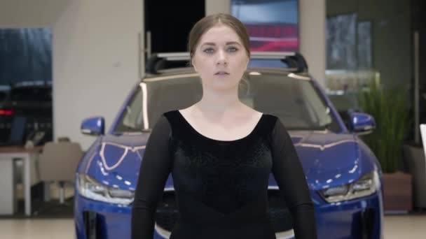 Bailarina de ballet caucásica sonriente balanceándose en puntillas. Elegante joven morena bailando en concesionario de coches delante del vehículo. Cámara bajando de bailarines cara a pies. Elegancia, automóviles . — Vídeos de Stock