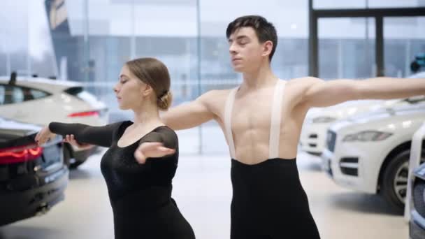 Bailarines de ballet caucásicos profesionales haciendo movimientos con las manos, mirando a la cámara y sonriendo. Hombre y mujer confiados bailando danza clásica en concesionario de coches. Industria automotriz, arte, elegancia . — Vídeo de stock