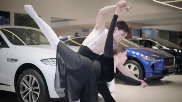 Kamera efter unga kaukasiska balettdansare som dansar i bilhandeln. Stilig man och vacker kvinna utför klassisk dans i showroom. Konst, elegans, bilindustri. — Stockvideo