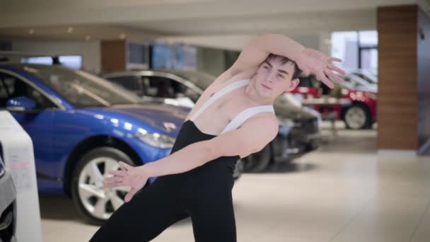 Retrato del joven hombre caucásico confiado bailando ballet en concesionario de automóviles. Bailarina de ballet profesional doblando y girando delante de los coches. Arte, industria automotriz, belleza . — Vídeos de Stock