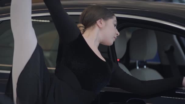 Bájos kaukázusi női balett táncos, lábával felfelé az autókereskedésben. Profi balerina portré a fekete autó mellett. Autóipar, művészet, szépség, elegancia. — Stock videók