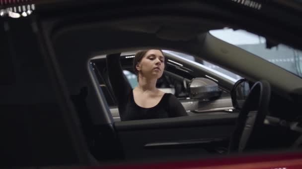 Graciösa kaukasiska ballerina dansar i bilhandlare. Porträtt av ung professionell balettdansare som gör klassiska drag i auto showroom. Skjuter genom bilsalongen. Långsamma rörelser. — Stockvideo