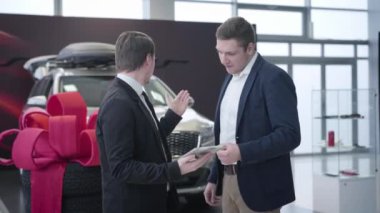İki beyaz adam araba galerisinde dikilip konuşuyorlar. Galeride elinde tablet ve danışman müşterisi olan bir araba satıcısı. Ticaret, otomobil endüstrisi, iş.