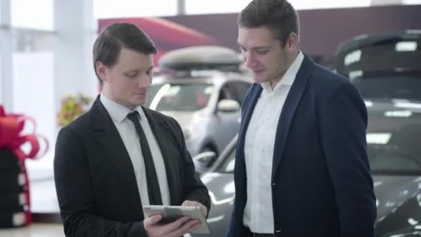 Retrato de negociante de carro masculino caucasiano e cliente usando tablet e olhando ao redor no showroom do carro. Dois homens em ternos de negócios discutindo a compra de automóvel na concessionária de carros. Negócios, estilo de vida . — Vídeo de Stock