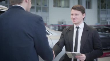 Esmer, beyaz, mavi gözlü tüccar portresi müşterilerin elini sıkıyor ve konuşuyor. Galeride müşteriye servis yapan genç bir araba satıcısı. Araba işi, ticaret, bayilik..