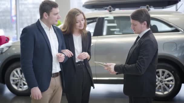 Běloch v obleku mluví s mladým párem v prodejně aut. Úspěšná rodina kupuje nové vozidlo v showroomu. Úspěch, obchod, automobilový průmysl. — Stock video