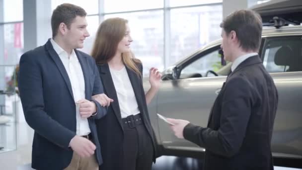 Jonge Kaukasische succesvolle paar selecteren van nieuw voertuig in de auto-dealer. Lachende man en vrouw aan het praten met een vertegenwoordiger in de showroom. Bedrijven, auto-industrie, levensstijl. — Stockvideo