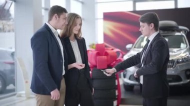 Yeni araba galerisinden anahtarlarını alan mutlu, beyaz bir ailenin yan görüntüsü. Kocası tüccarla tokalaşırken gülümseyen kadın torbacıdan araba anahtarlarını alıyor. Başarı, iş.