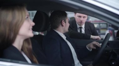 Yakışıklı, beyaz bir adamın karısıyla arabada oturup torbacıyla konuşmasının yan görüntüsü. Gülümseyen adam ve kadın araba galerisinde yeni bir araç seçiyor. Otomobil endüstrisi, iş dünyası, satın alma.