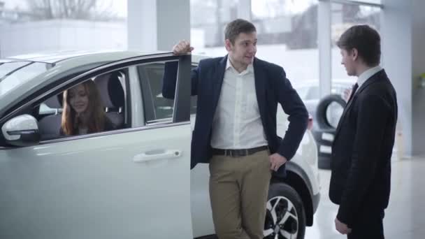 Šťastná kavkazská mladá žena sedící v autě, zatímco její manžel mluví s dealerem. Úspěšný pár kupující vozidlo v showroomu. Automobilový průmysl, automobilový průmysl. — Stock video
