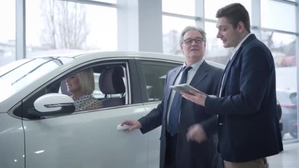 Bella donna bionda caucasica matura seduta in auto come suo marito anziano che parla con il rivenditore di auto nello showroom. Uomo d'affari anziano di successo l'acquisto di nuovo veicolo per il suo coniuge . — Video Stock