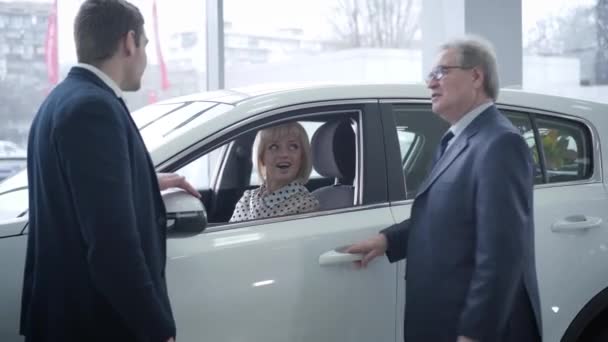 Two Caucasian men standing next to new car in dealership as beautiful mature woman sitting inside. Three people in dealership discussing purchase. Business, lifestyle, success. — 비디오