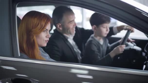 Vista lateral de la hermosa pelirroja caucásica mujer sentada en el salón de coches con la familia. Enfoque cambios a marido e hijo sentados en el asiento del conductor y sosteniendo el volante. Felicidad, compra, éxito . — Vídeos de Stock