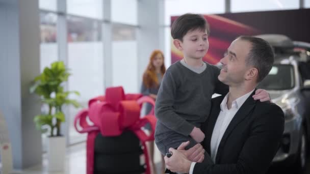 Portrait of confident Caucasian businessman holding son on hands and giving him car keys. Cheerful blurred woman making victory gesture at the background. Family in car dealership. — Stock Video