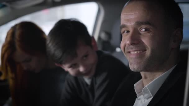 Gros plan portrait d'un homme caucasien satisfait regardant la caméra et souriant alors que sa famille est assise à l'arrière-plan. Famille dans le showroom automobile assis dans le véhicule. Affaires, joie, succès . — Video