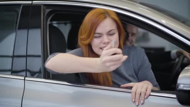 Retrato de una hermosa pelirroja caucásica sosteniendo las llaves del coche y haciendo un gesto de victoria. Alegre madre sentada en un automóvil nuevo con marido e hijo. Industria automotriz, alegría, felicidad . — Vídeos de Stock