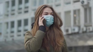 Koruyucu maskeli genç bir kadın şehirde duruyor ve telefonda konuşuyor. Esmer kız öksürüyor. Küresel tehlike, bulaşıcı virüs, salgın.