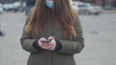 Şehir caddesinde koruyucu maske takan tanınmayan esmer kadın. Telefonda daktilo eden genç bir kadın. Coronavirus, izolasyon, küresel tehlike.