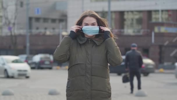 Střední záběr mladé brunetky, jak stojí na ulici a nasazuje si ochrannou masku. Úzkostlivá žena, která se chrání před virem epidemie. Pandemie, infekce, globální katastrofa. — Stock video