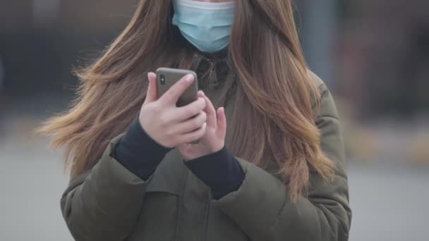 Närbild av oigenkännlig ung kvinna i skyddande mask med hjälp av smartphone. Brunett kvinna står på stadens gata läser nyheter om coronavirus. Medicin, sjukvård, fara. — Stockvideo