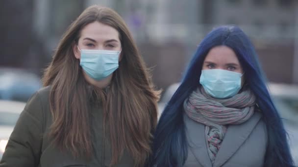 Portrét dvou žen v ochranných maskách, které se dívají na kameru, jak stojí na ulici. Mladé kamarádky používají ochranné masky na ochranu proti koronaviru. Pandemie, epidemie, nebezpečí. — Stock video
