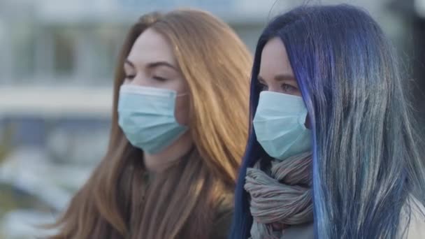 Vista lateral de cerca de dos mujeres ansiosas con máscaras protectoras mirando hacia otro lado. Mujeres en la calle de la ciudad. El enfoque cambia de chica de pelo azul a mujer morena. Peligro global, peligro, virus . — Vídeos de Stock