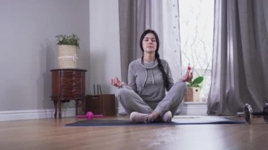 Kamera, evde meditasyon yapan kendine güvenen esmer beyaz kadına yaklaşıyor. Yoga minderinde gözleri kapalı ve bacak bacak üstüne atmış genç bir kadın. Huzur, yaşam tarzı, rahatlama..