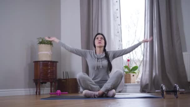 Longo tiro de morena caucasiana mulher sentada no tapete de ioga levantar as mãos e colocá-los juntos. Meditação de mulher forte confiante dentro de casa. Exercício, ioga, desporto . — Vídeo de Stock