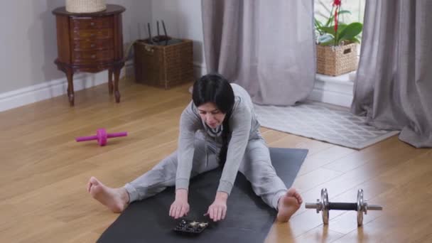 Vue du dessus de la jeune belle femme caucasienne étirant vers des bonbons comme assis sur le tapis de yoga. Bonne femme brune faisant de l'exercice et mangeant des bonbons à la maison. Mode de vie, sport, yoga . — Video