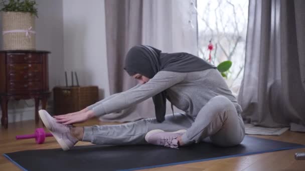 Vista lateral de la delgada mujer musulmana inclinándose hacia adelante en la esterilla de yoga tratando de llegar de puntillas con las manos. Mujer joven y deportiva practicando yoga en casa. Motivación, fitness, estilo de vida . — Vídeos de Stock