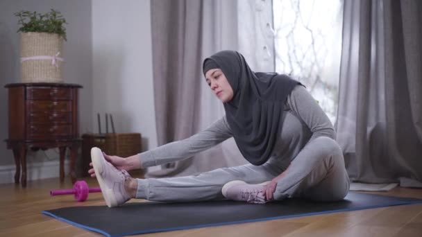 Mulher muçulmana jovem em sportswear e hijab flexão no tapete de ioga tocando dedos do pé. Mulher forte bonita praticando ioga em casa. Motivação, fitness, estilo de vida . — Vídeo de Stock