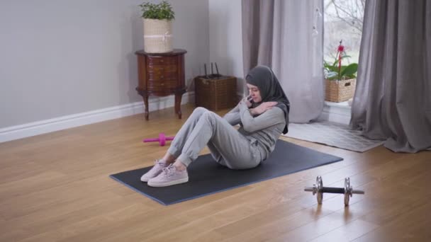 Jonge sterke moslimvrouw pompt de pers naar binnen. Slanke sportieve vrouw in hijab en sportkleding versterkt de spieren thuis. Sport, fitness, levensstijl. Bovenaanzicht, kleine kans. — Stockvideo