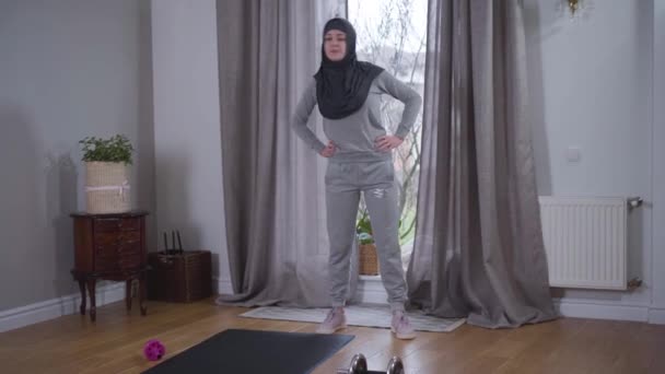 Retrato de una joven musulmana en ropa deportiva de pie en casa y haciendo a un lado. Mujer hermosa fuerte en el hiyab practicando en el interior. Entrenamiento, ejercicio, estilo de vida . — Vídeo de stock