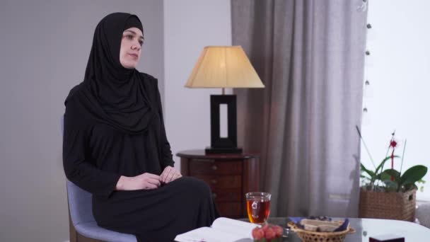 Ritratto di triste donna musulmana in abiti neri seduta a casa da sola. Bella donna sconvolta in hijab guardando fuori dalla finestra e pensando. Stile di vita, solitudine, frustrazione . — Video Stock