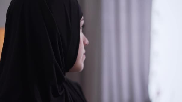 Närbild ansikte vacker upprörd kvinna i hijab tittar ut genom fönstret hemma. Porträtt av sorgsen muslimsk kvinna i traditionella kläder. Ensamhet, depression, sorg. — Stockvideo
