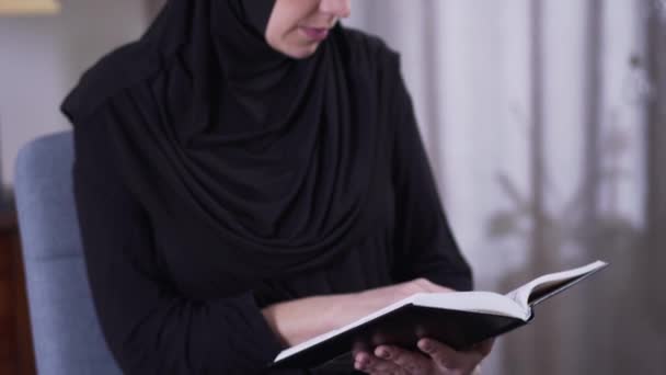 Câmera movendo-se de livro para mulher muçulmana focada lendo. Mulher moderna inteligente em roupa tradicional descansando em casa. Hobby, inteligência, estilo de vida . — Vídeo de Stock