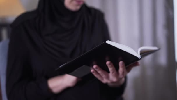 Primer plano del libro en manos de mujeres musulmanas. Mujer borrosa en el hiyab negro tradicional leyendo en el fondo. Estilo de vida, hobby, literatura . — Vídeos de Stock