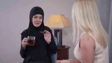 Mutlu Müslüman kadın portresi. Kurabiyeyle çay içen ve sarışın beyaz bir arkadaşla konuşan. Farklı kültürlerden iki modern kadının arkadaşlığı. İletişim, topluluk, sosyal.