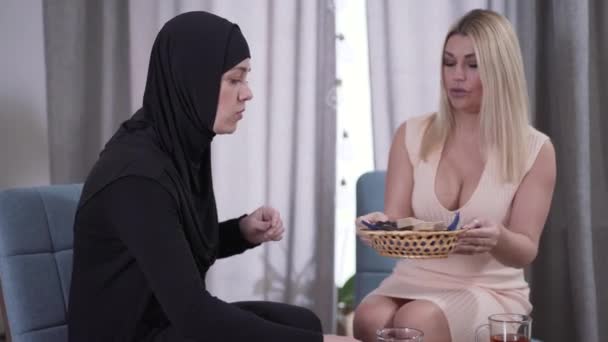 Blond positif Femme caucasienne traitant ami musulman avec des bonbons. Deux femmes de cultures différentes qui boivent du thé et parlent à l'intérieur. Amitié, diversité, différence culturelle, mode de vie . — Video