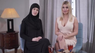 Farklı kültürlerden iki kadının portresi kameraya bakıyor ve gülümsüyor. Beyaz tenli sarışın kadın ve geleneksel tesettürlü Müslüman kadın içeride oturuyor. Çeşitlilik, kültürel farklılık