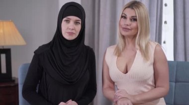 Beyaz ve Müslüman kadınlar kameraya bakıp gülümsüyor. Samimi elbiseli sarışın modern kadın ve siyah tesettürlü Müslüman kadın içeride poz veriyor. Kültürel çeşitlilik, hoşgörü.