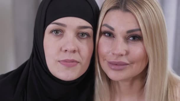 Visi ravvicinati di donna musulmana in hijab e donna bionda caucasica. Due donne diverse di culture diverse che guardano la macchina fotografica e sorridono. Tolleranza, coesistenza, stile di vita . — Video Stock