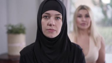 Kameraya bakan tesettürlü genç Müslüman kadın portresi. Arka planda duran bulanık, modern beyaz kadın. Güzellik, kültürel çeşitlilik, yaşam tarzı.